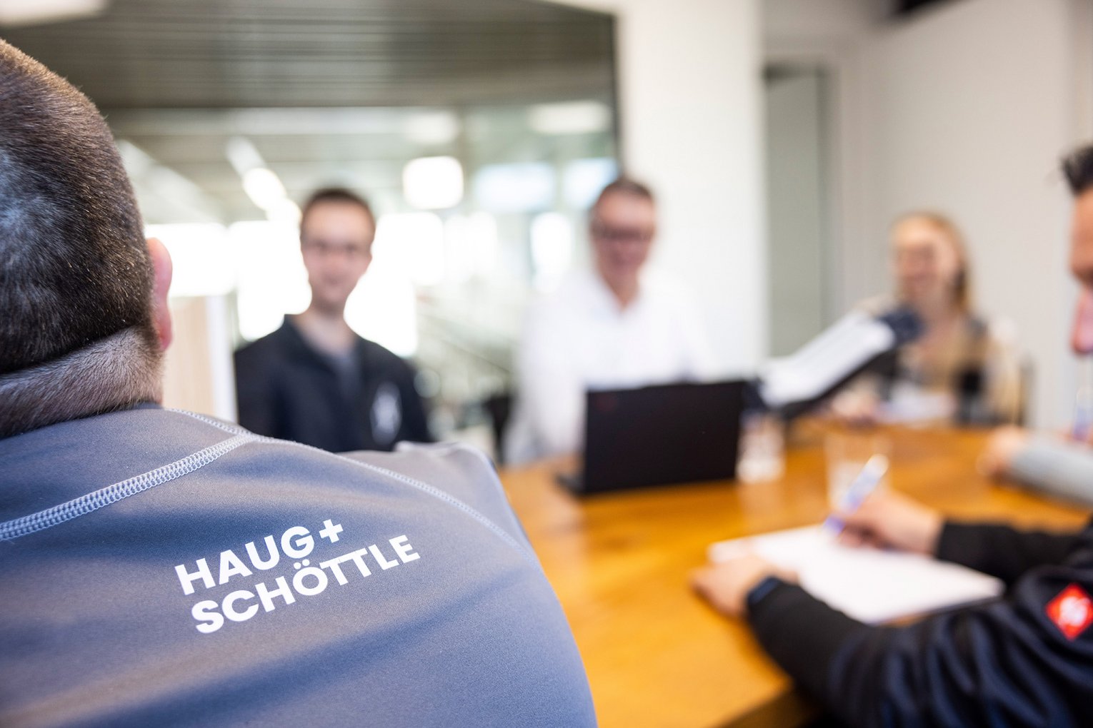 Meeting bei HAUG + SCHÖTTLE Das Team von HAUG + SCHÖTTLE hält ein Meeting an einem großen Holztisch, im Hintergrund sitzen Personen mit Laptops